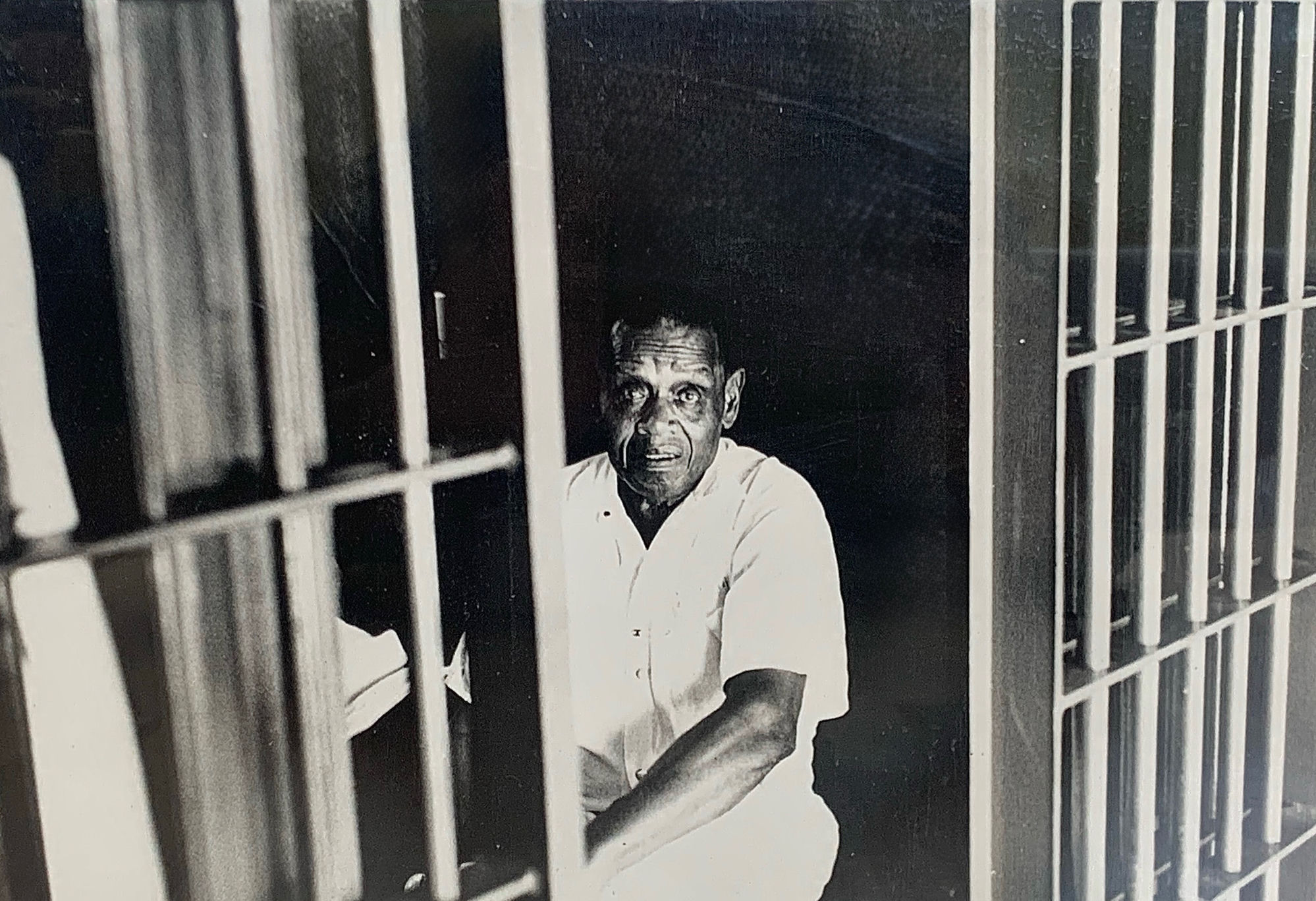 Frank Jones seated behind the cell bars in a white shirt, looking up toward the camera.